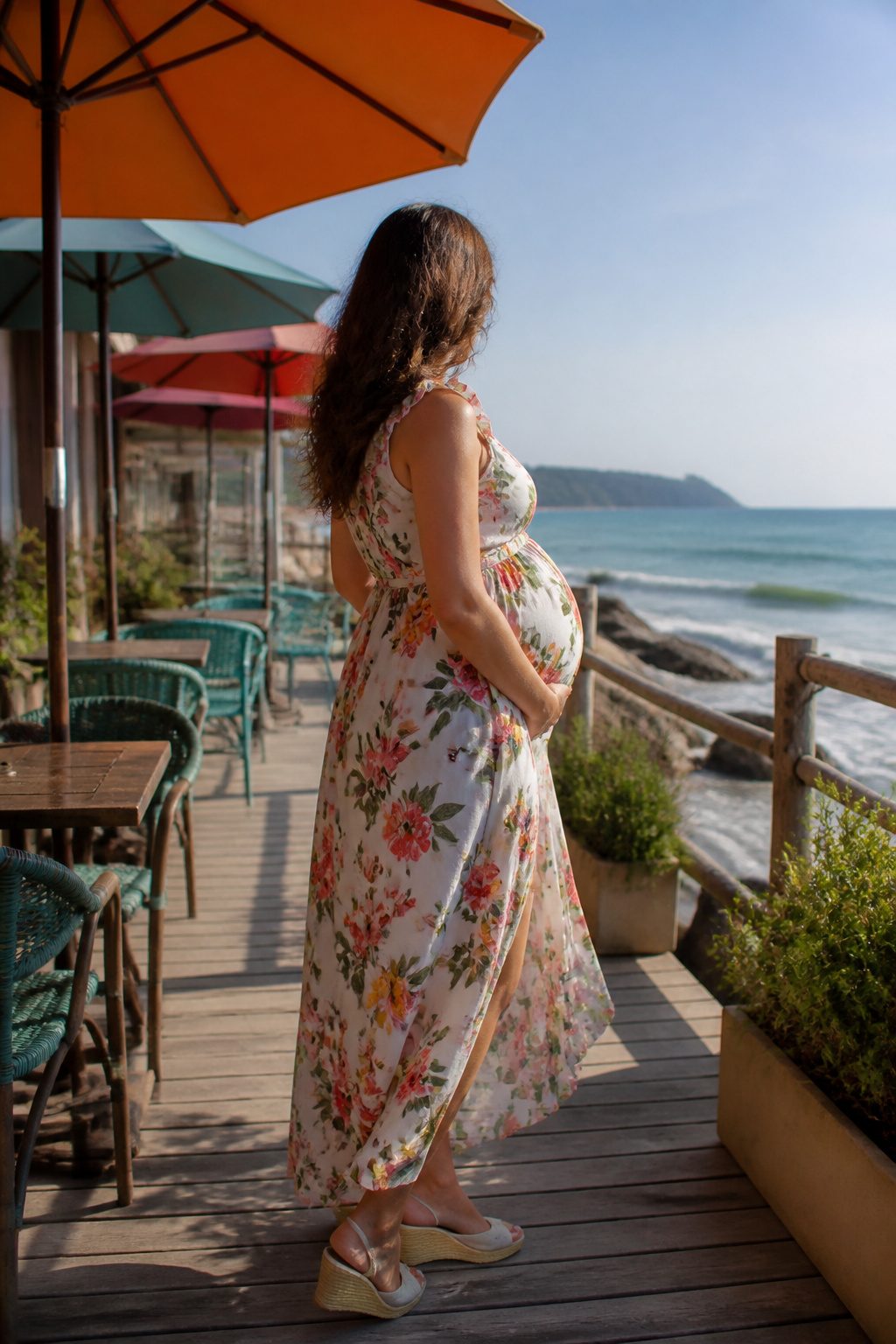 Expectant woman in a sleeveless floral maternity dress by the beach.